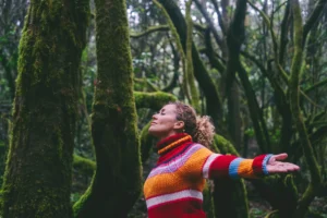 Mujer con los brazos abiertos y los ojos cerrados en medio del bosque, conectando con la naturaleza y su mundo interior.