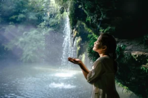 Niña con los ojos cerrados y las manos abiertas bajo el agua de una cascada, disfrutando de un momento de conexión con la naturaleza.