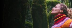 Mujer con los brazos abiertos y los ojos cerrados en medio del bosque, conectando con la naturaleza y su mundo interior.