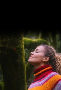 Mujer con los brazos abiertos y los ojos cerrados en medio del bosque, conectando con la naturaleza y su mundo interior.