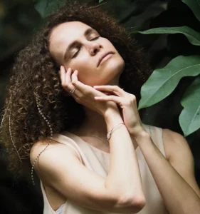 Mujer con los ojos cerrados y expresión de paz, sosteniendo su rostro con las manos en un entorno natural.