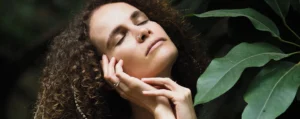 Mujer con los ojos cerrados y expresión de paz, sosteniendo su rostro con las manos en un entorno natural.