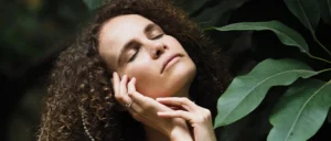 Mujer con los ojos cerrados y expresión de paz, sosteniendo su rostro con las manos en un entorno natural.