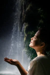Niña con los ojos cerrados y las manos abiertas bajo el agua de una cascada, disfrutando de un momento de conexión con la naturaleza.