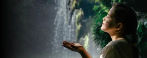 Niña con los ojos cerrados y las manos abiertas bajo el agua de una cascada, disfrutando de un momento de conexión con la naturaleza.