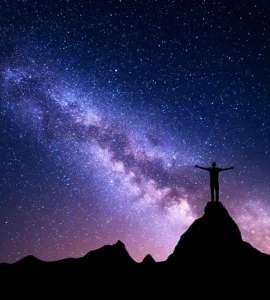 Silueta de un hombre con los brazos abiertos sobre una montaña, contemplando un cielo estrellado.