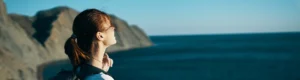 Mujer de perfil con los ojos cerrados frente al mar, de pie al borde de una montaña en un momento de conexión interior.